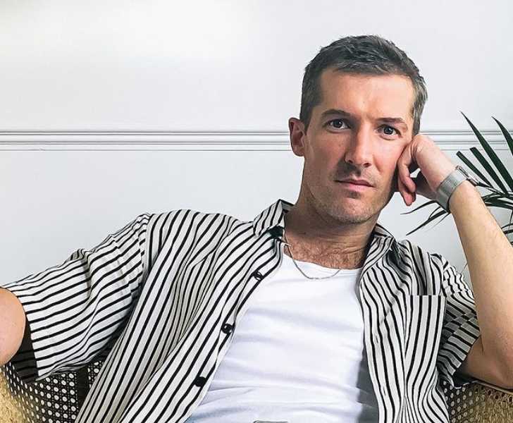 Picture of Gwilym Lee posing for a photo by putting his left hand on his left chick white themed picture with white wall on background